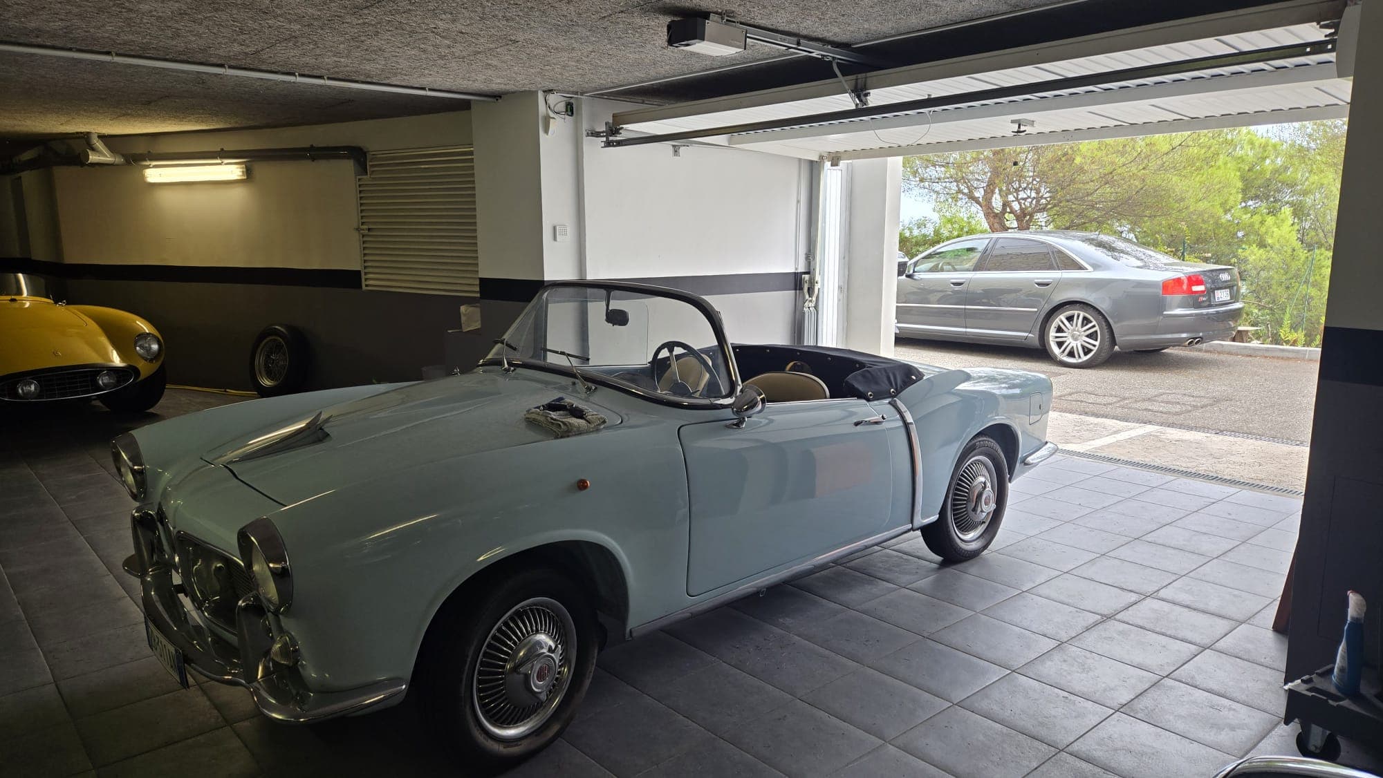 Light blue vintage convertible in a garage with a yellow sports car and open door.