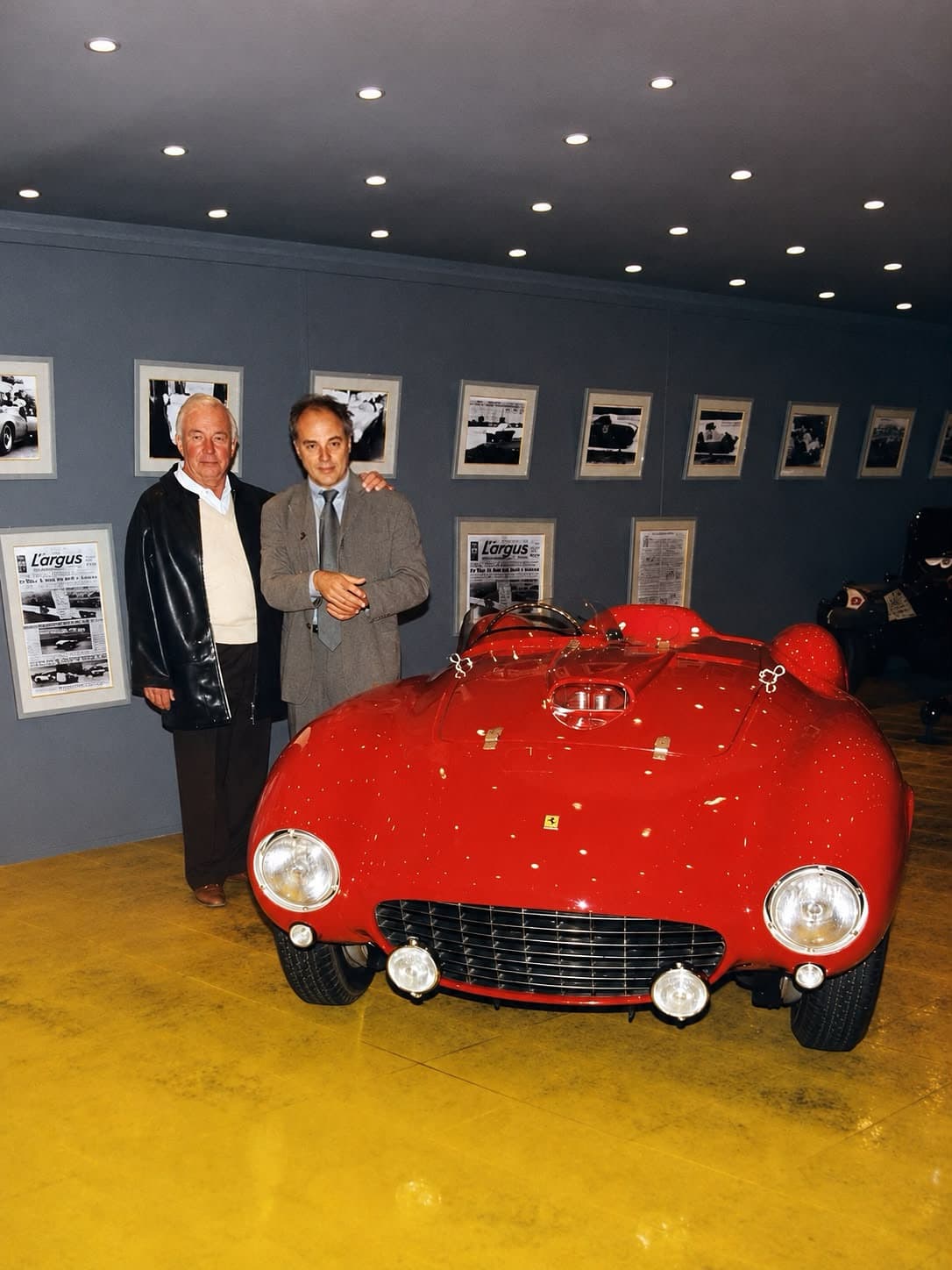 Two men standing behind a vintage red Ferrari in a gallery with framed photos.