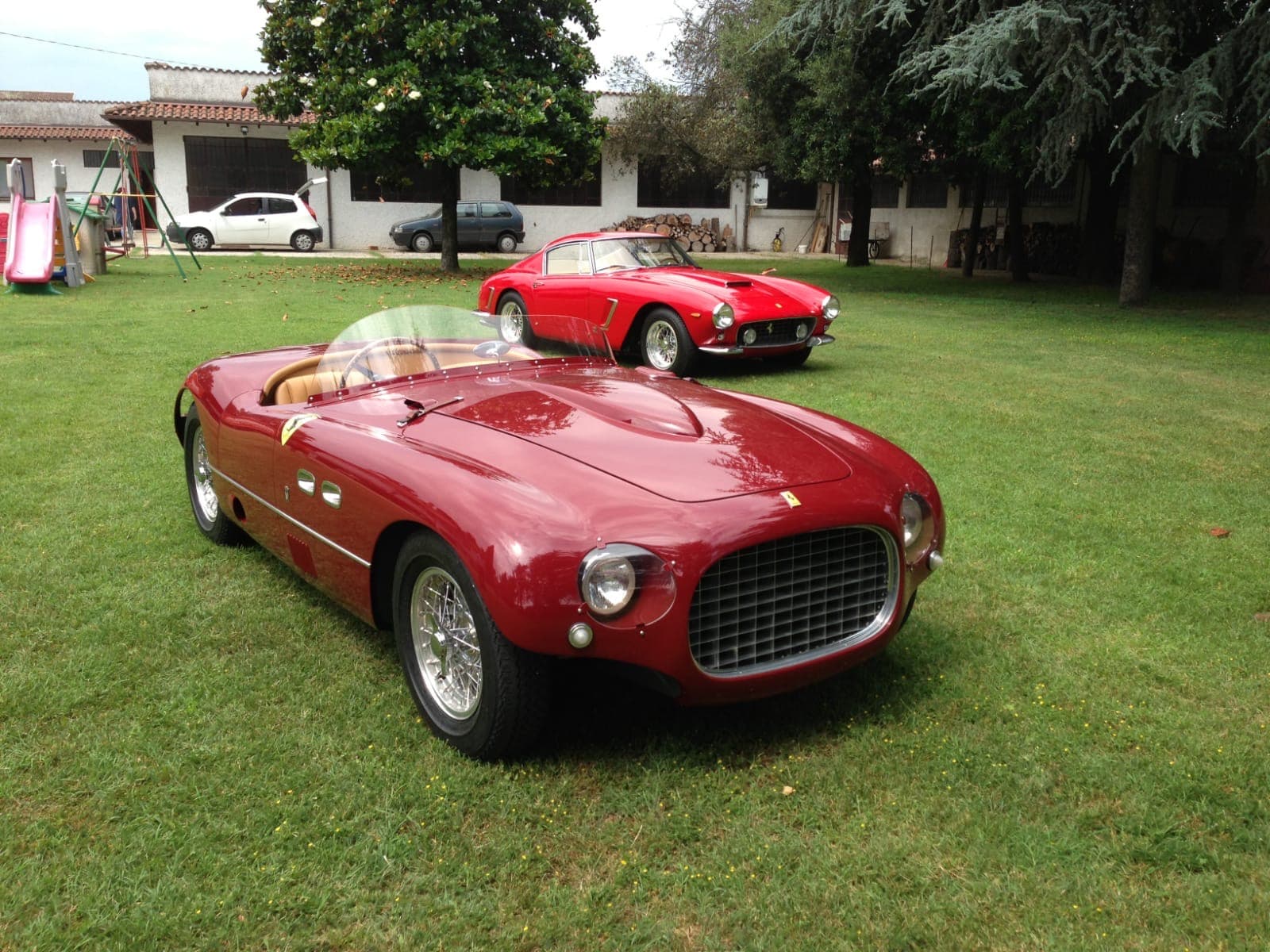 Two vintage red Ferraris, a roadster and a coupe, parked on a green lawn.