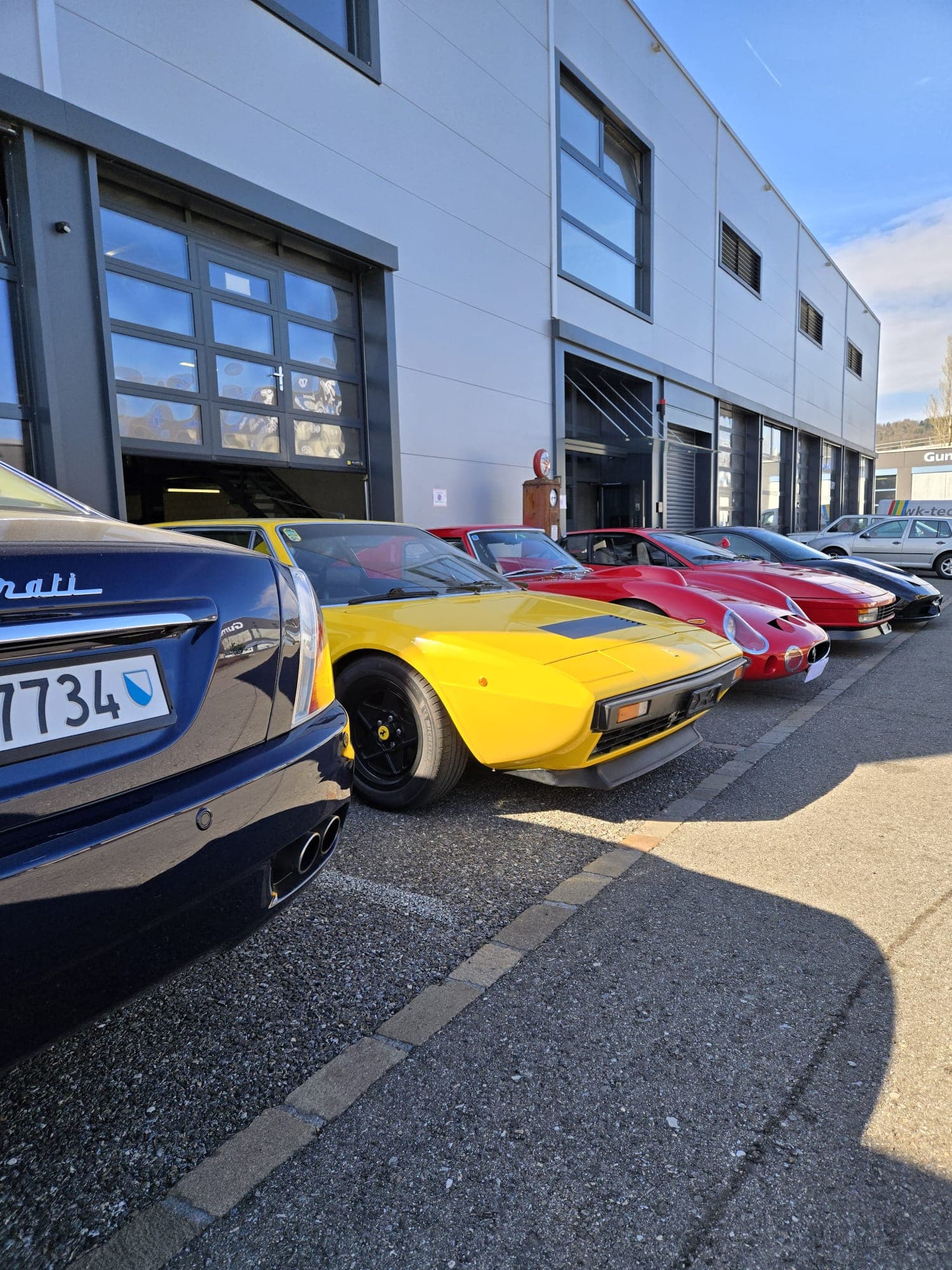 Blue Maserati and row of colorful Ferraris parked outside a modern grey industrial building.