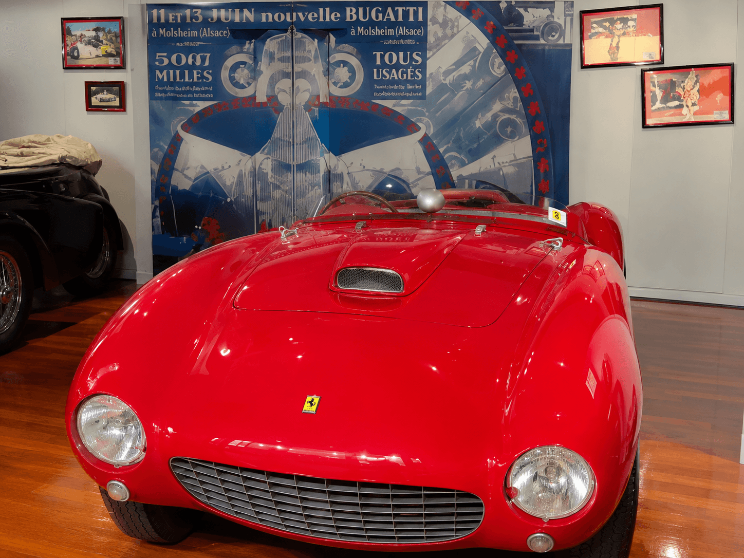 Red vintage Ferrari sports car displayed in a museum with a large Bugatti poster.