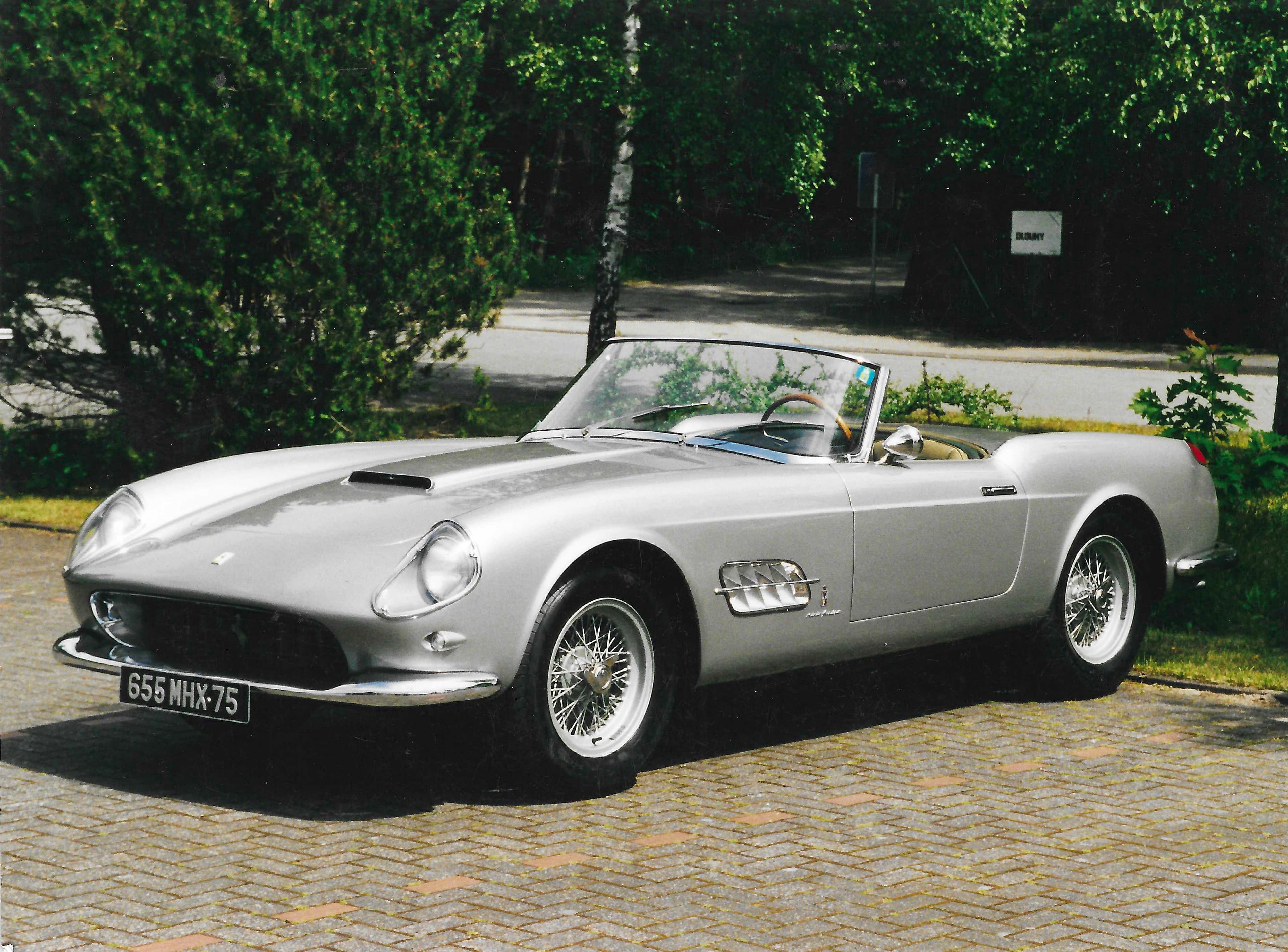 Classic silver Ferrari 250 GT California Spyder parked on a brick driveway with green trees.