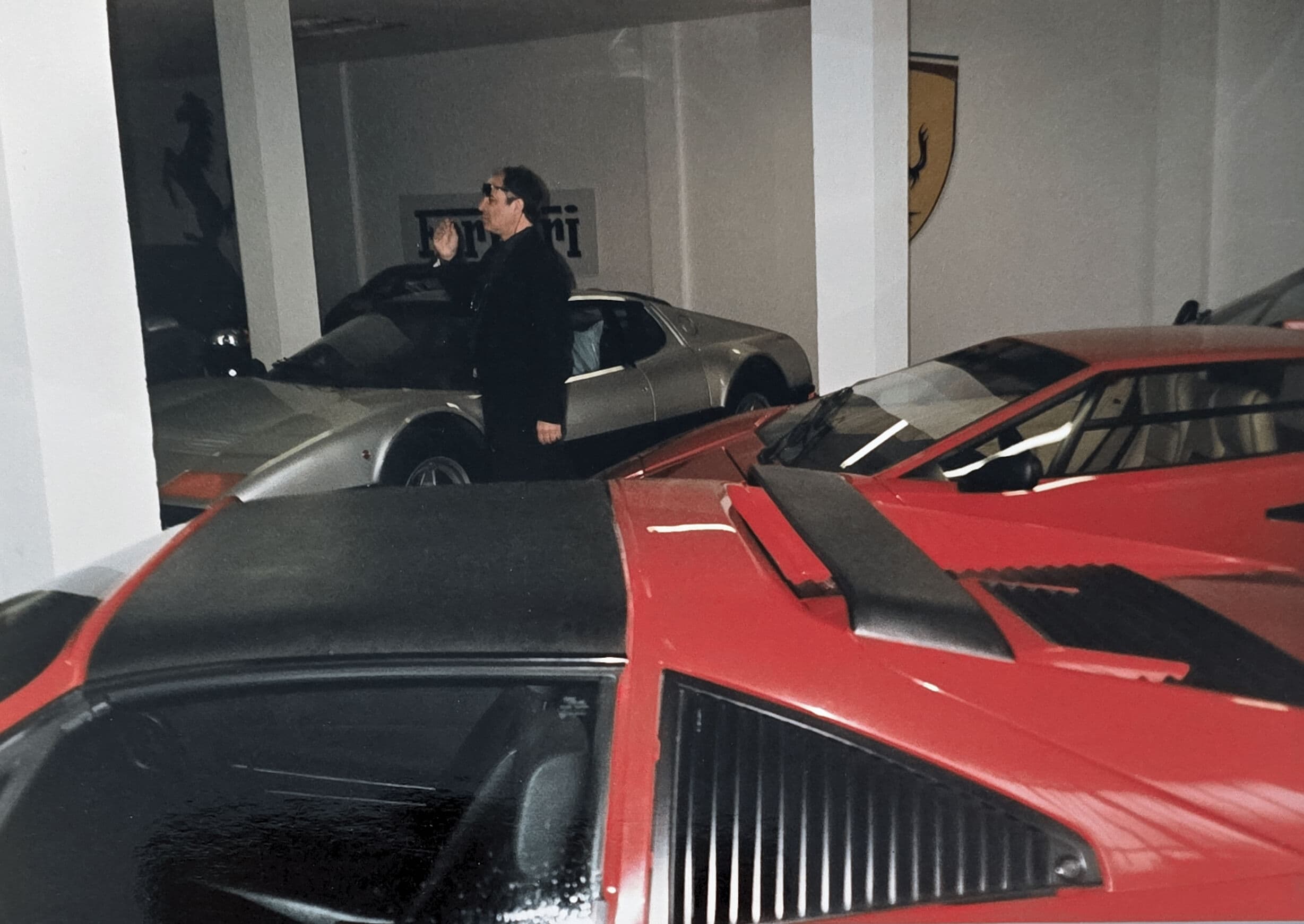 Man in black suit standing among vintage red and silver Ferrari sports cars.