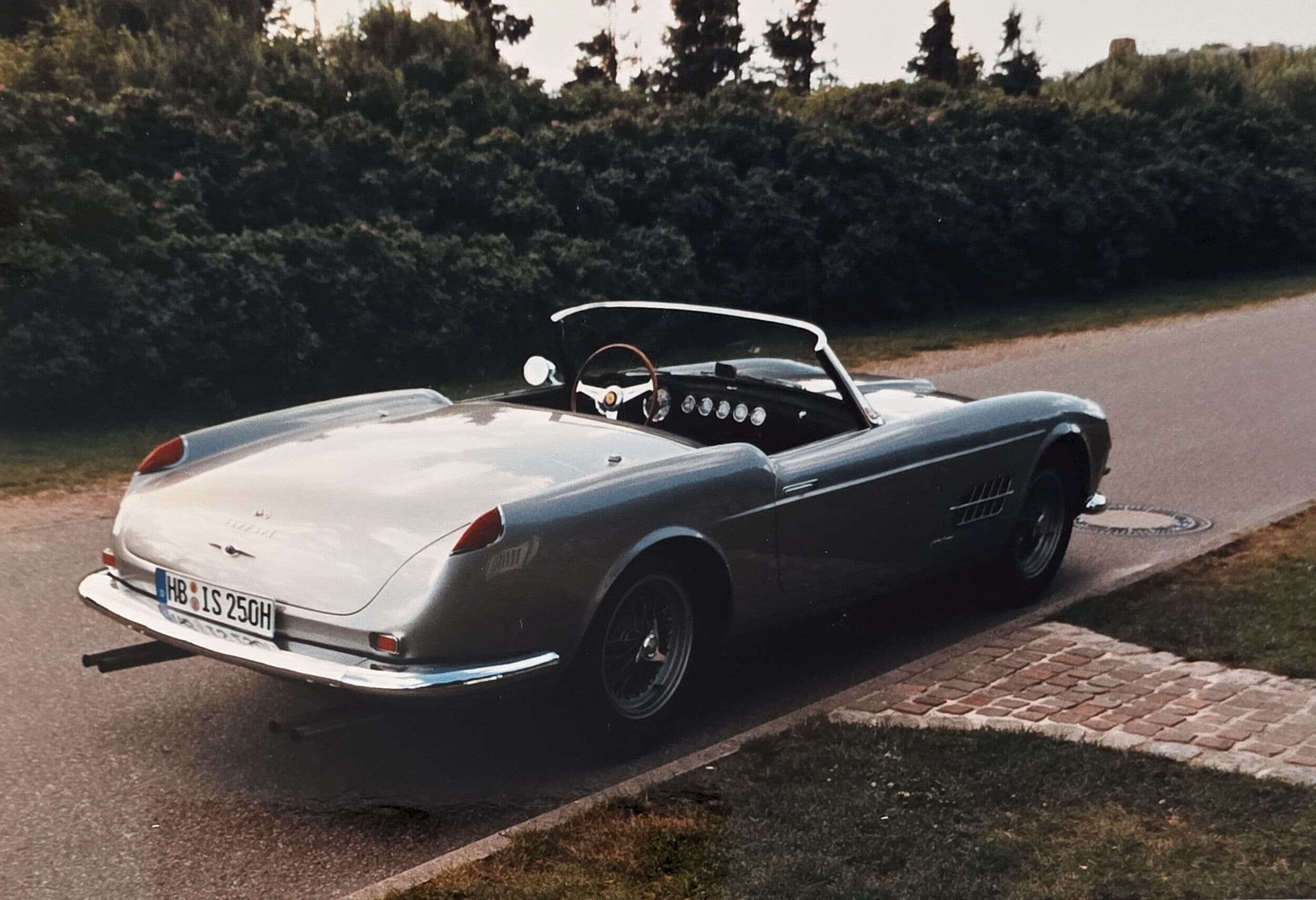 Classic silver Ferrari convertible parked on a roadside next to a lush green hedge.