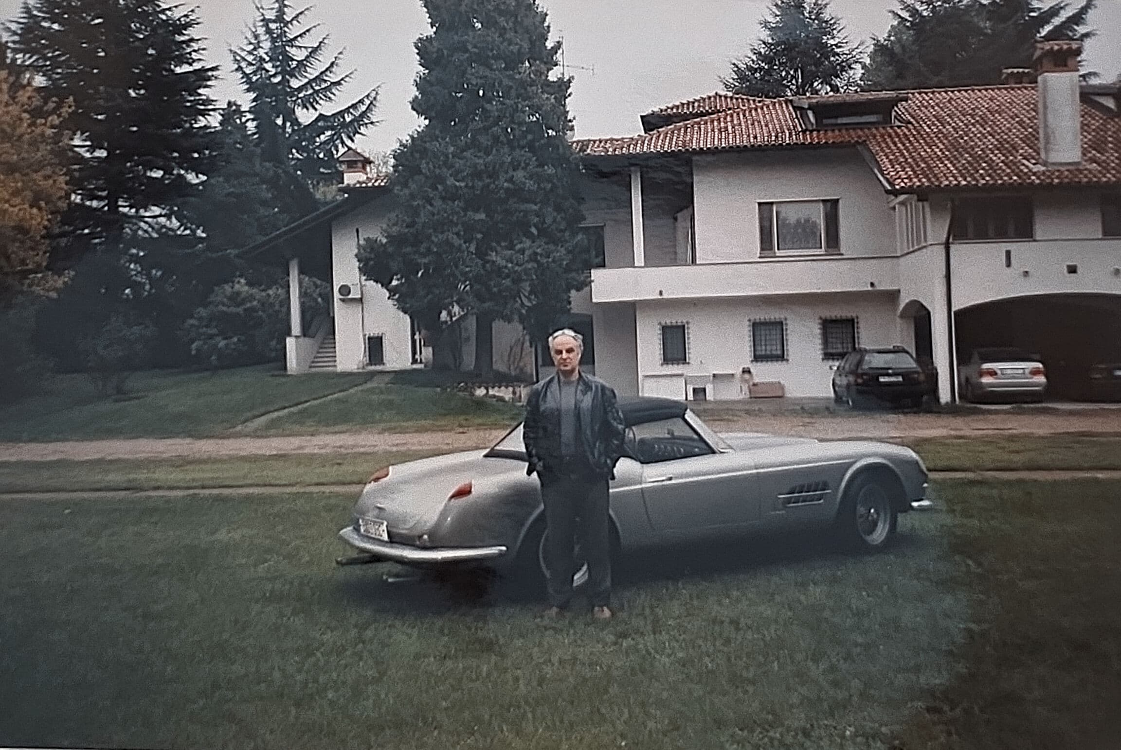 Man stands beside a silver vintage sports car in front of a large white villa.