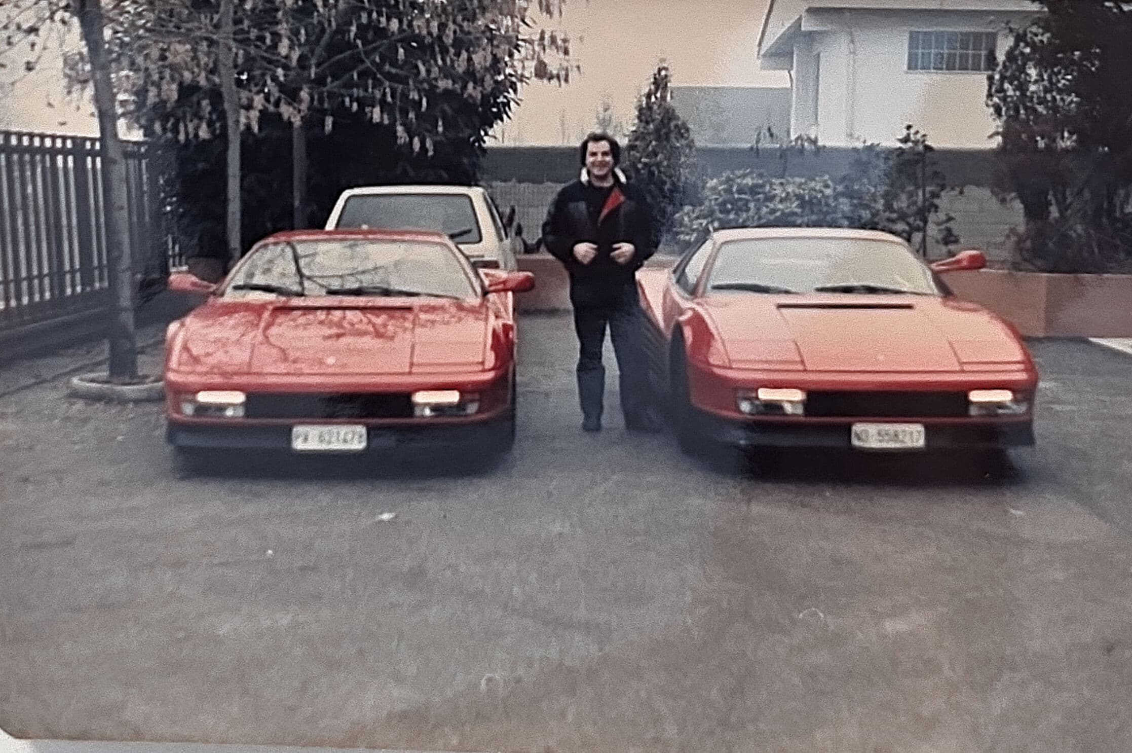 Man posing between two identical red Ferrari Testarossa sports cars parked in a driveway.