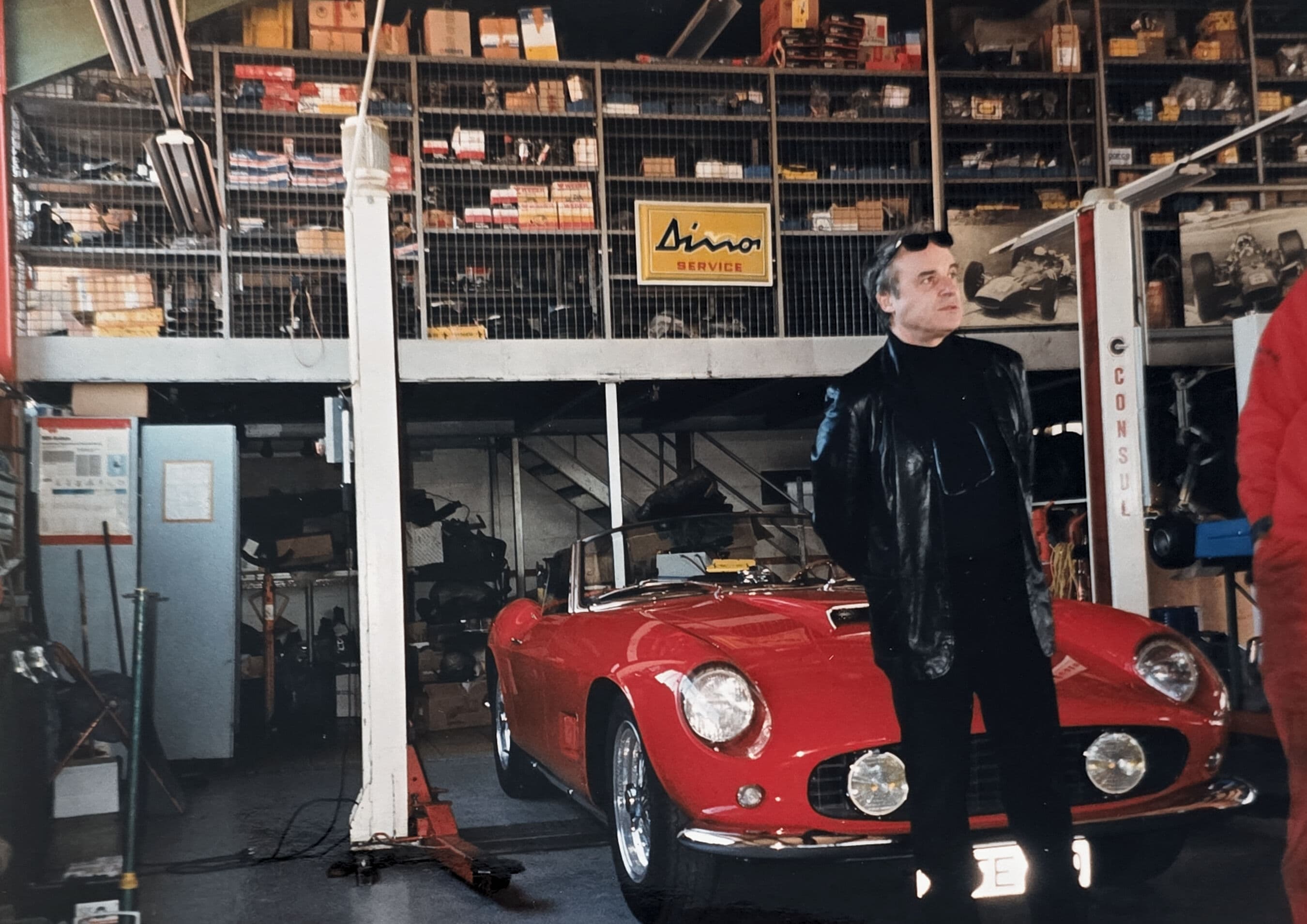 Man in black leather jacket stands before a classic red convertible in a vintage garage.