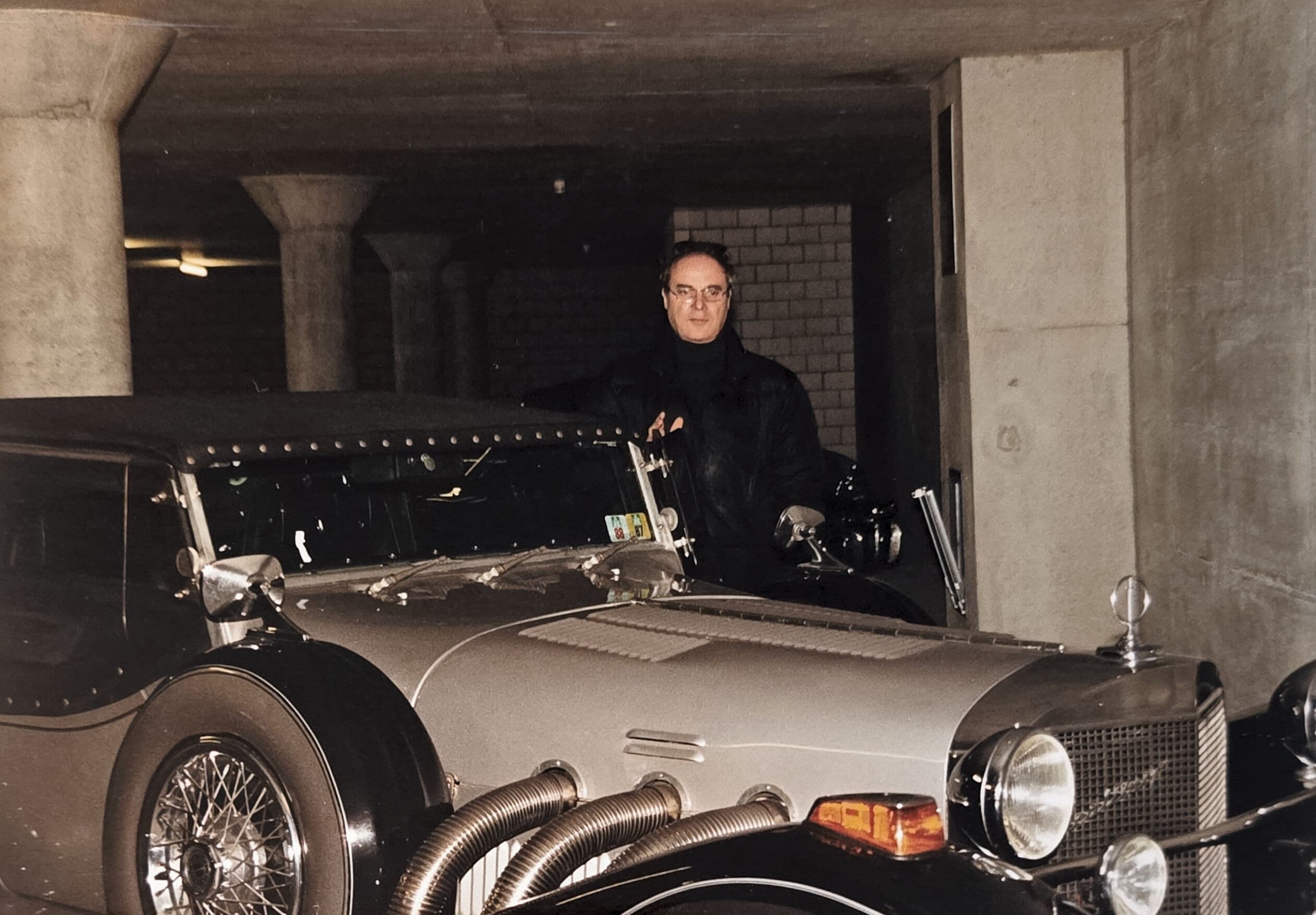 Man in glasses stands behind a vintage silver car with exposed exhaust pipes.