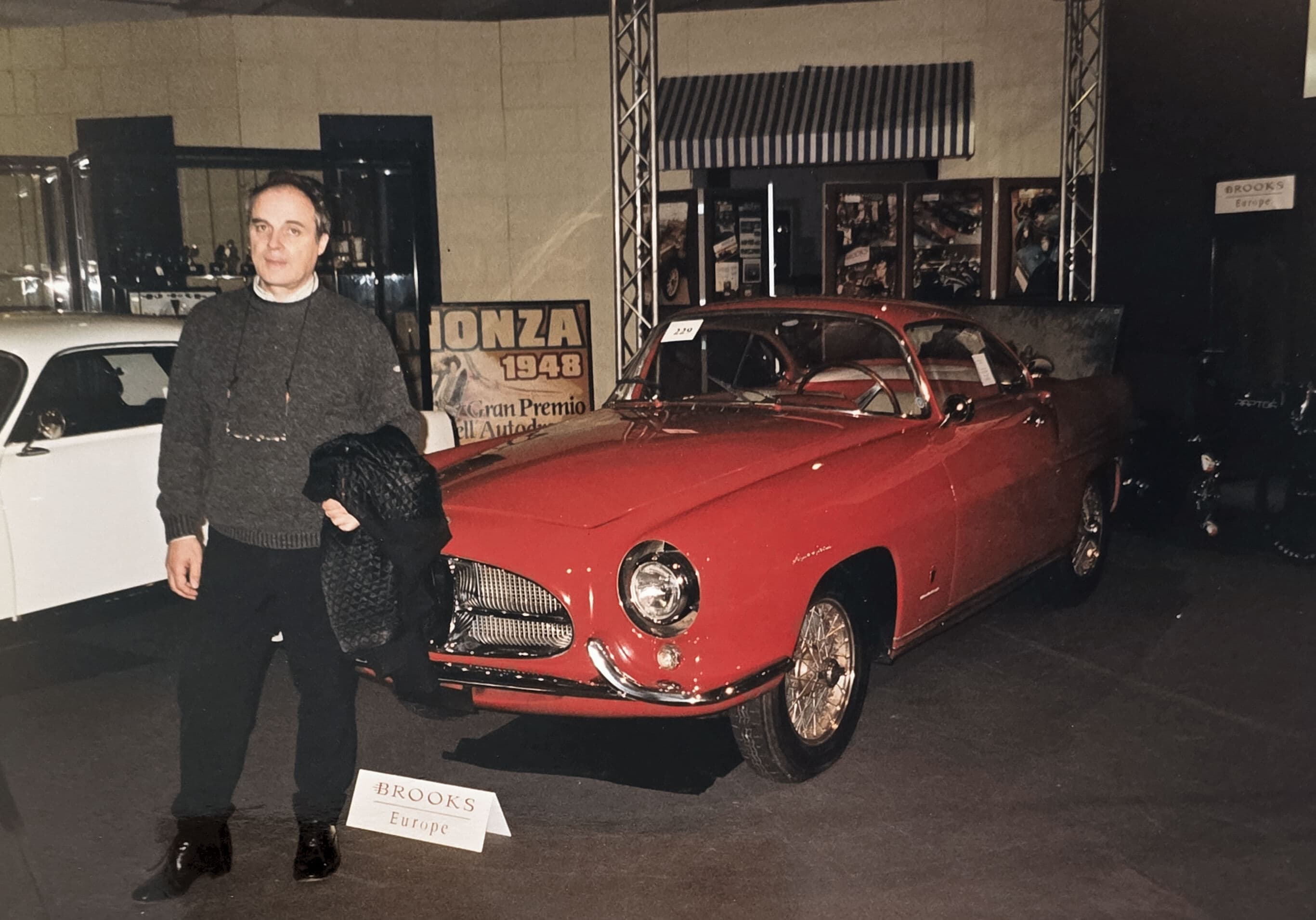 Man in grey sweater posing next to a vintage red sports car at an exhibition.
