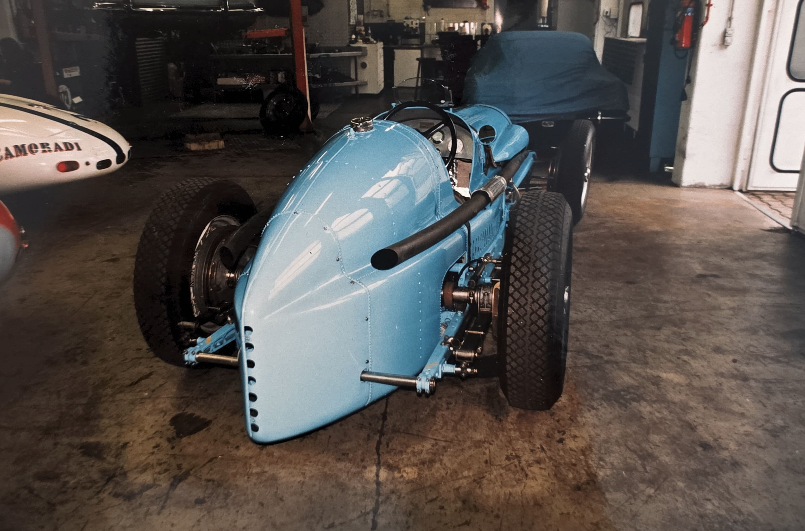 Vintage light blue open-wheel race car with a pointed tail parked in a garage workshop.