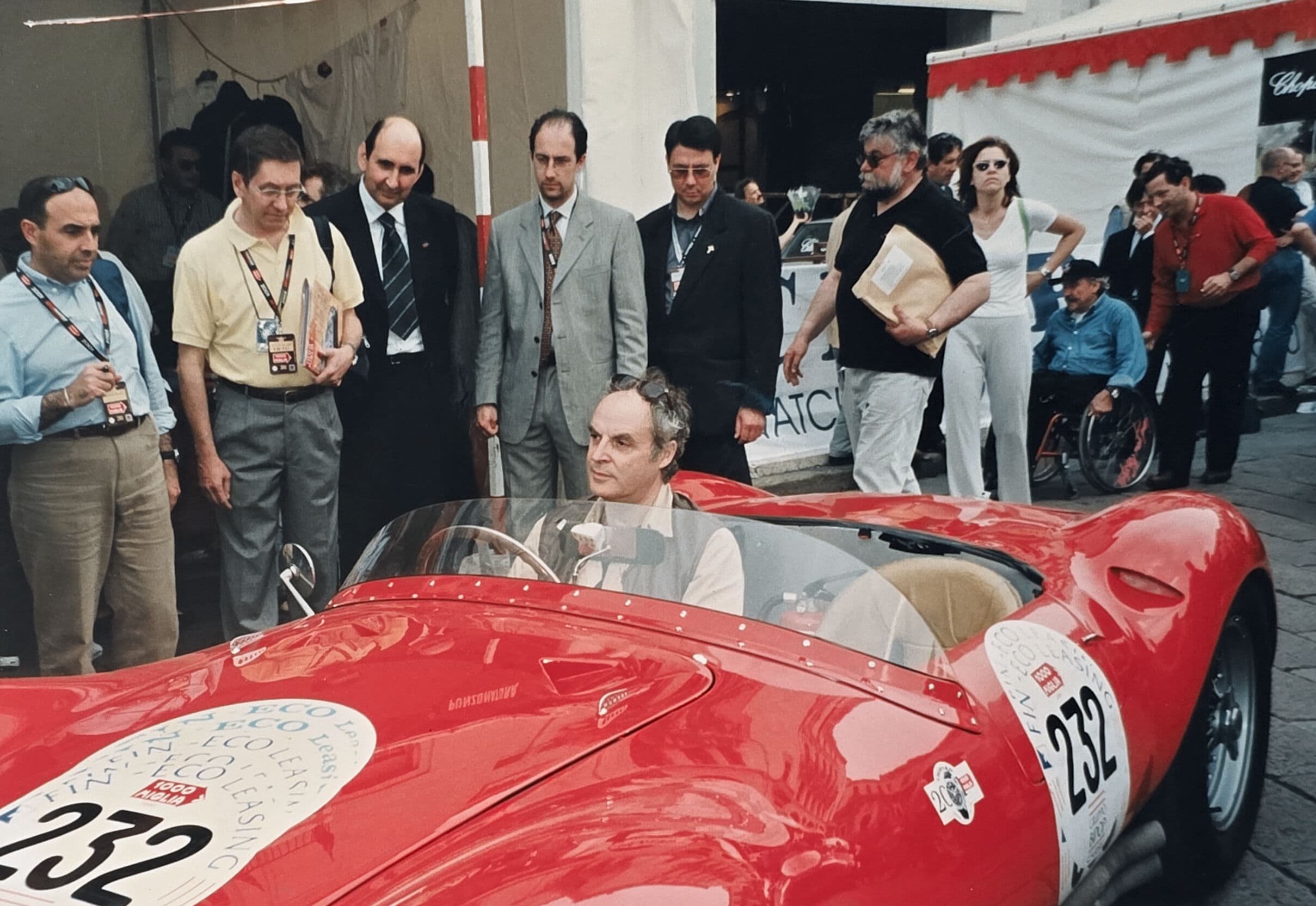 A man sits in a red vintage race car numbered 232, surrounded by onlookers.