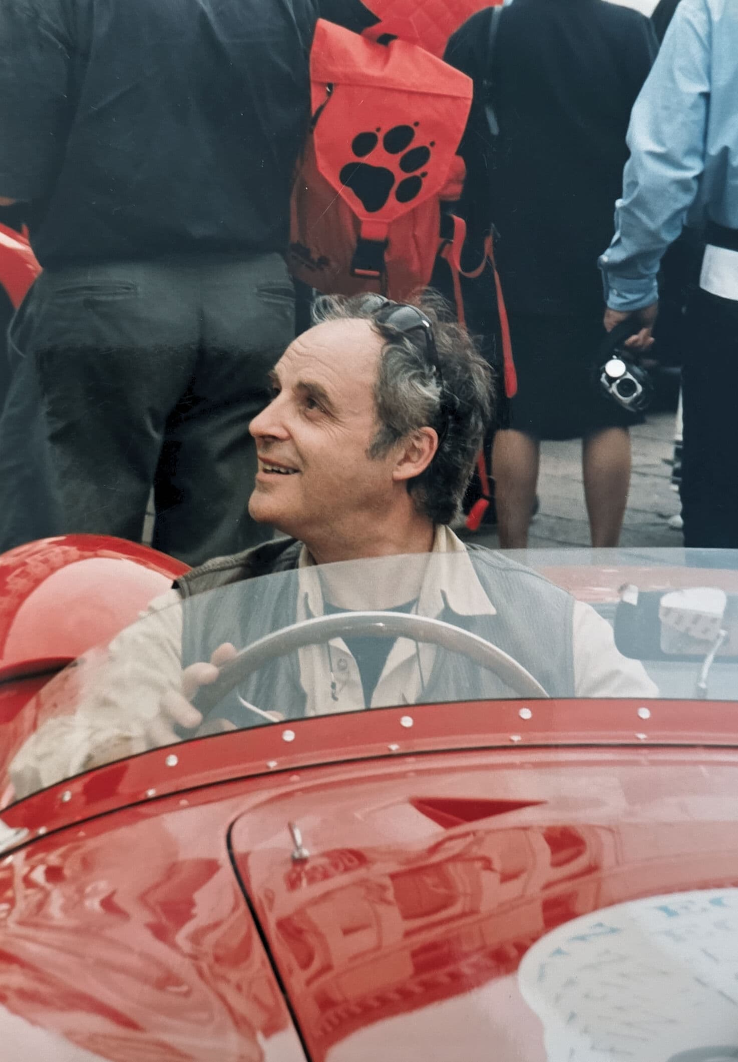 A smiling man sits in a vintage red open-top car surrounded by a crowd.