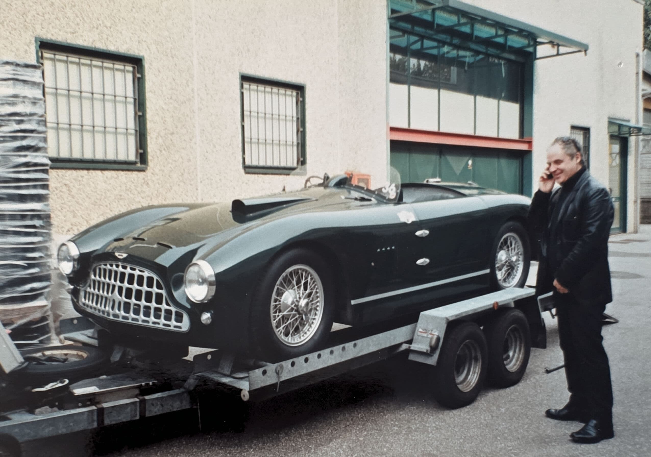 Vintage dark green sports car on a trailer beside a man talking on a phone.