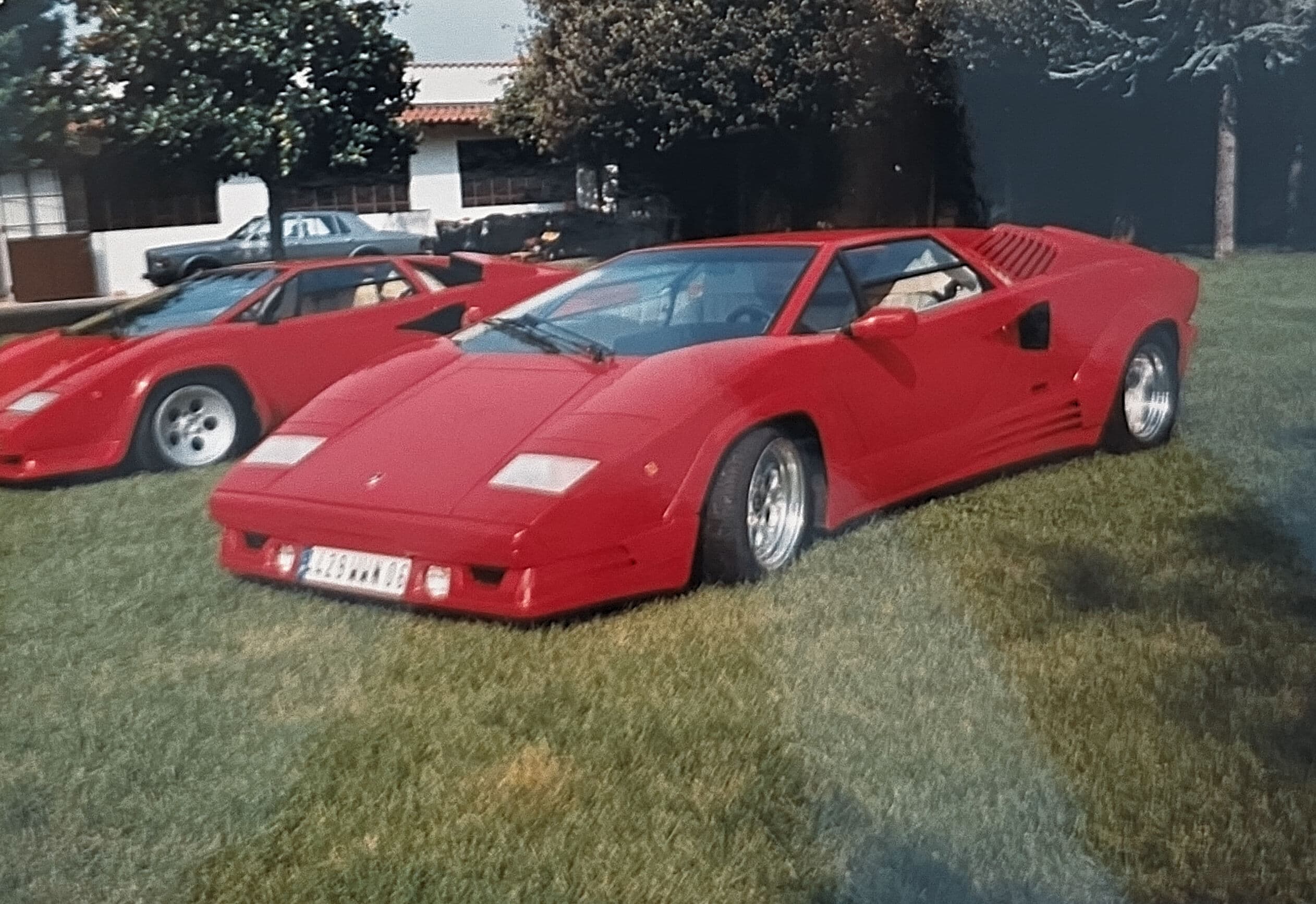 Two red Lamborghini Countach supercars parked on a green lawn with trees in the background.