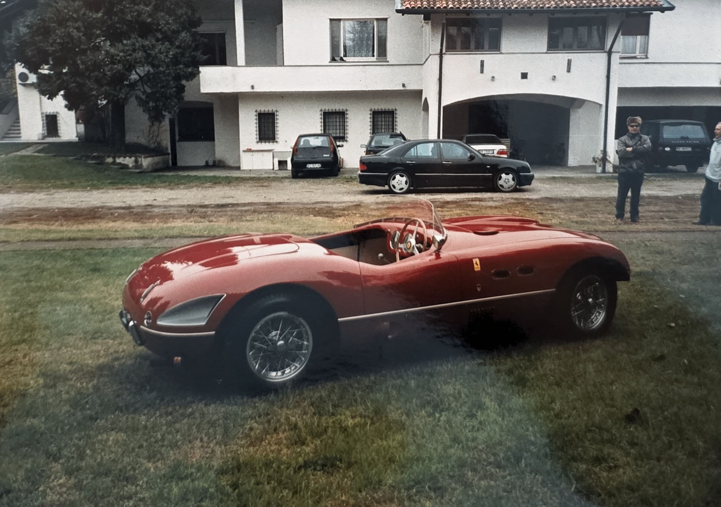 Classic red Ferrari open-top sports car parked on grass before a large white building.