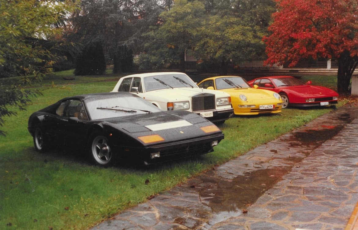 Black Ferrari, white Rolls-Royce, yellow Porsche, and red Ferrari parked on a wet lawn.