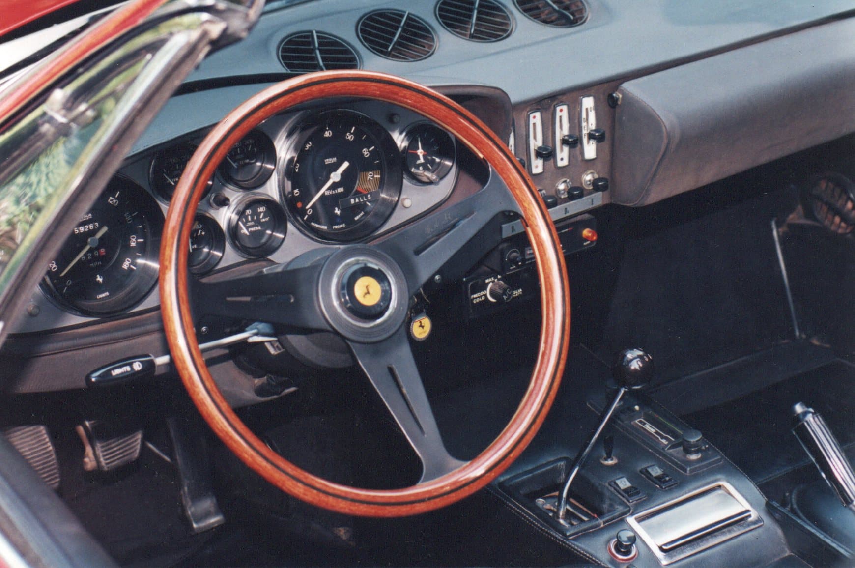 Classic Ferrari interior with a wooden steering wheel, analog gauges, and a gated manual shifter.