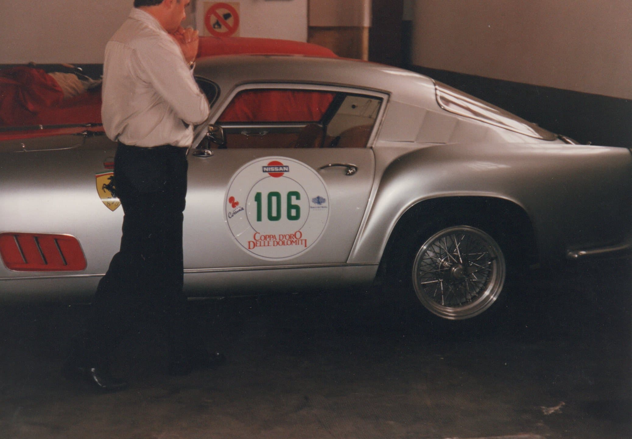 Man standing beside a silver vintage Ferrari race car with number 106 decal.