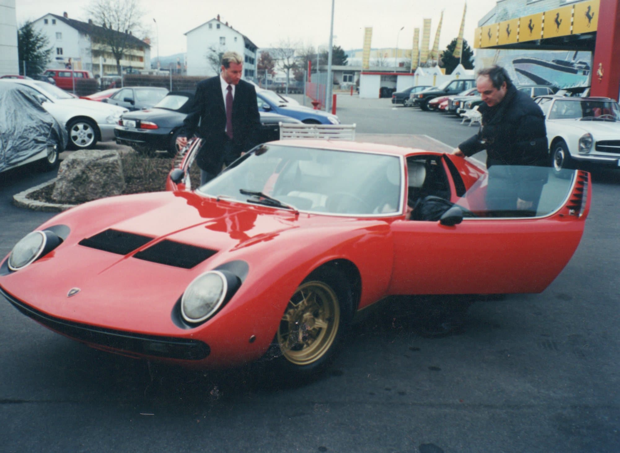 Red vintage Lamborghini Miura in a lot with two men standing by the open door.