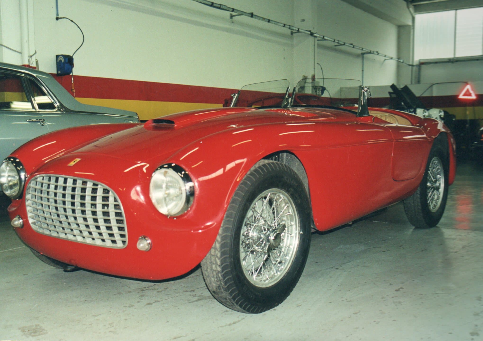 Vintage red Ferrari 166 MM Barchetta with wire wheels parked in a garage workshop.
