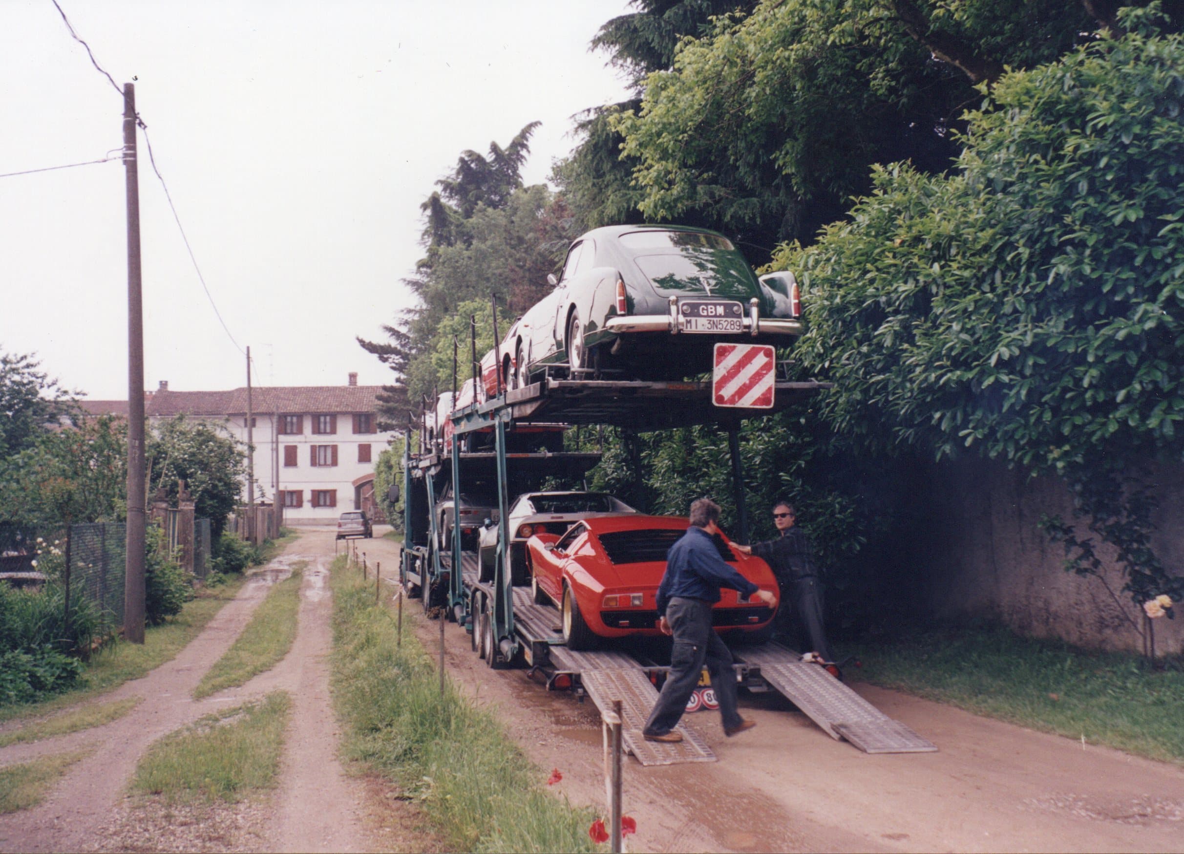 Vintage sports cars being loaded onto a transporter truck on a narrow rural dirt road.