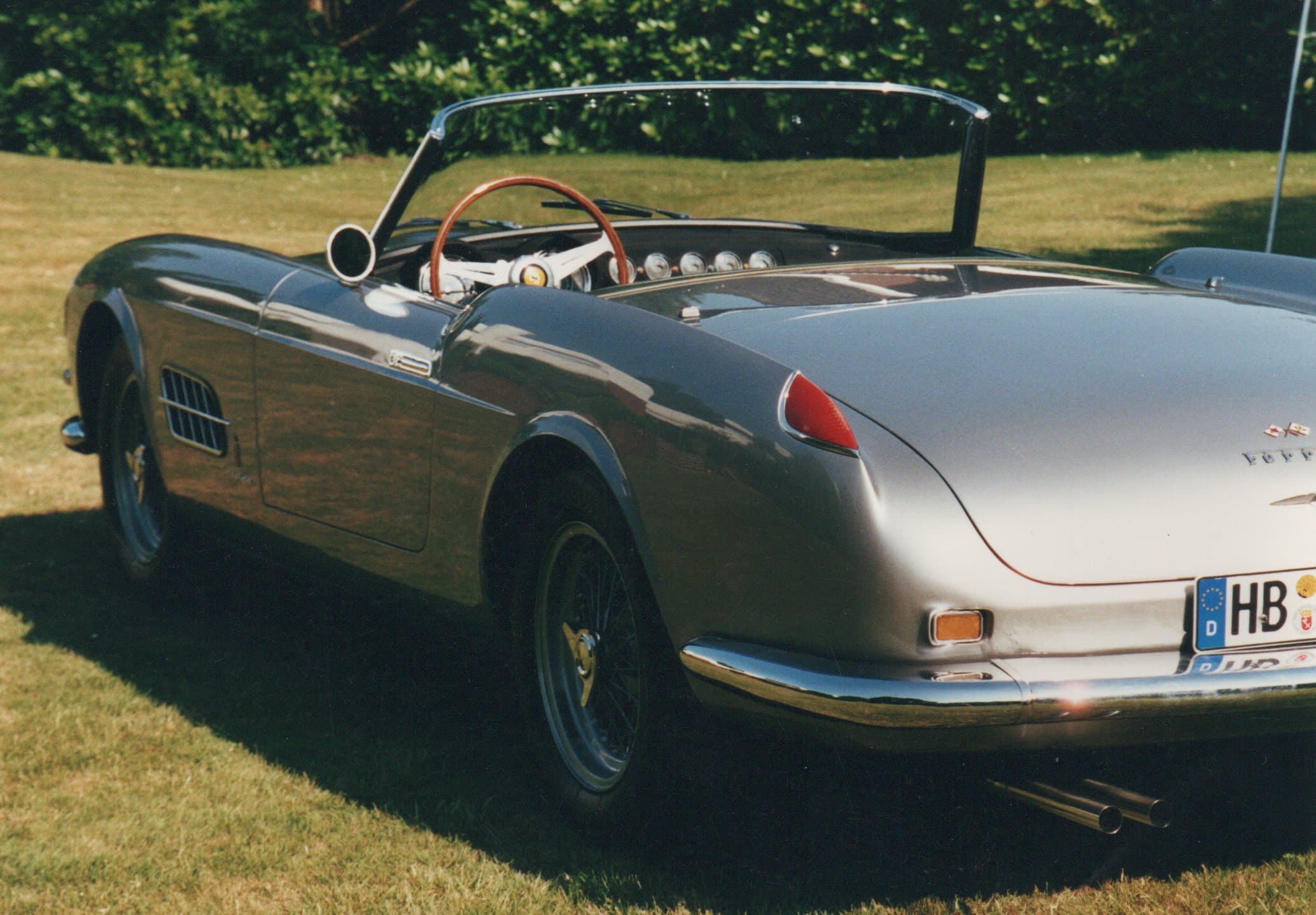 Classic silver Ferrari convertible with a wooden steering wheel parked on a green lawn.