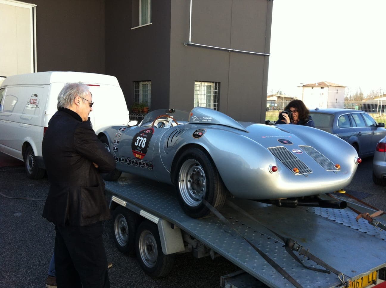 Silver vintage racing car number 378 secured on a trailer, observed by a man and photographer.
